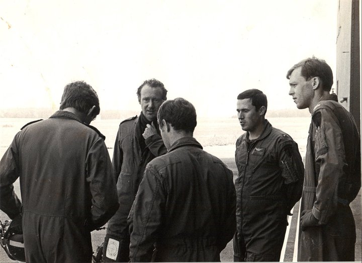 A black and white photo. A group of men in flight suits stand in a circle talking. Bob is pictured on the far right.