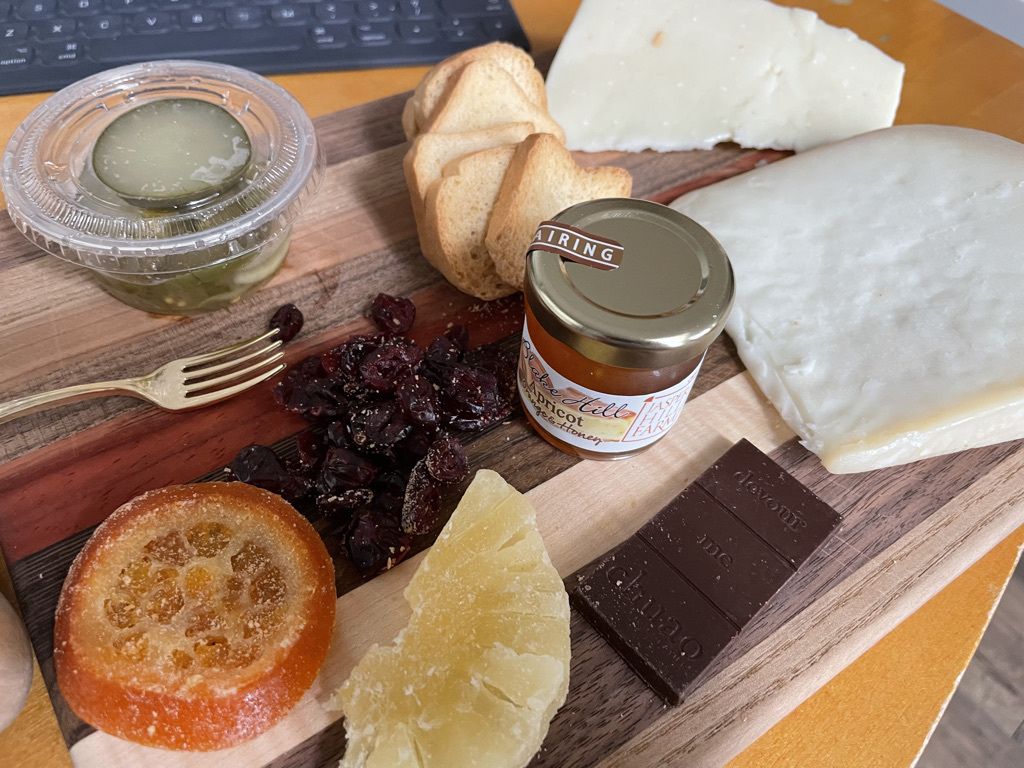 a cheese board with 2 types of cheese, mini toast squares, jam, pickles, candied fruit, and chocolate