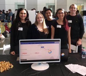 A group of five students stands behind their expo table. The table contains a monitor displaying a visual user interface for an application.