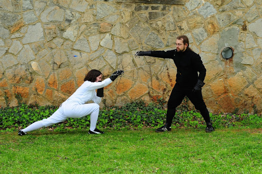 Two people (in fencing gear with holding swords) practice fencing.