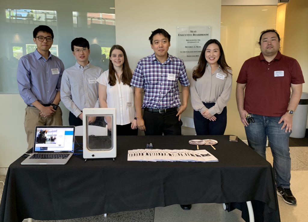 Students stand behind their expo table; the table contains a laptop displaying a software demo, a catdoor used for the demo, brochures for the application, and images of cat, dog, and wolf faces,