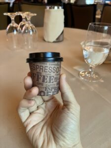 A hand holding a 2-ounce paper takeout espresso cup with plastic lid
